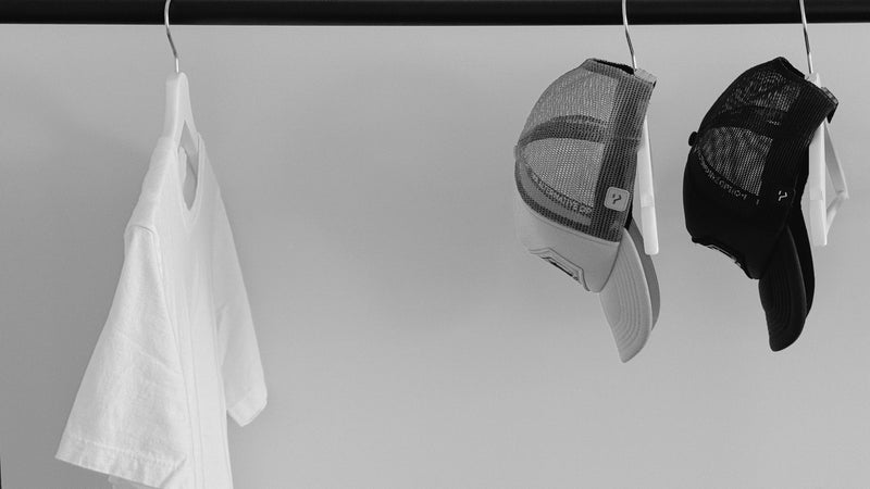 Black and white image of The Alternative Option’s Lowgo Tee and two Other Trucker Hats hanging on a clothing rack, captured in a minimalist setting to reflect fewer, better essentials.