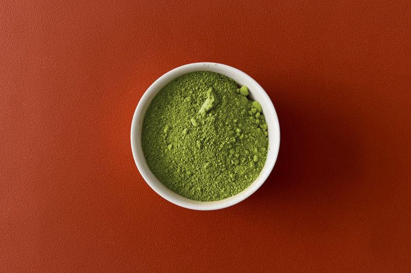 A small white bowl of vibrant green matcha powder on a rich, red-orange surface—capturing the simplicity and purity at the heart of traditional Japanese tea culture.