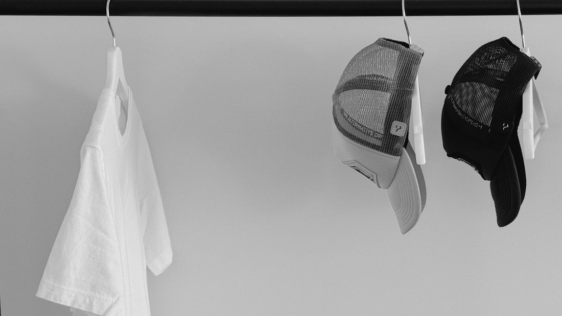 Black and white image of The Alternative Option’s Lowgo Tee and two Other Trucker Hats hanging on a clothing rack, captured in a minimalist setting to reflect fewer, better essentials.